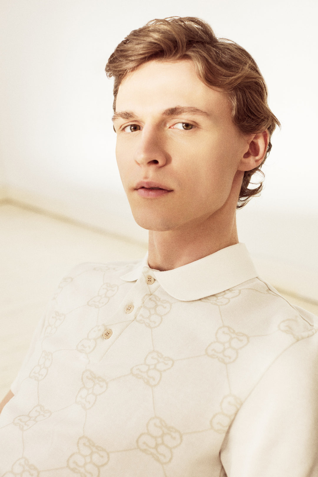 A close-up portrait of a young man with light brown hair wearing a cream-colored collared shirt with a subtle beige Gucci double-G monogram pattern.